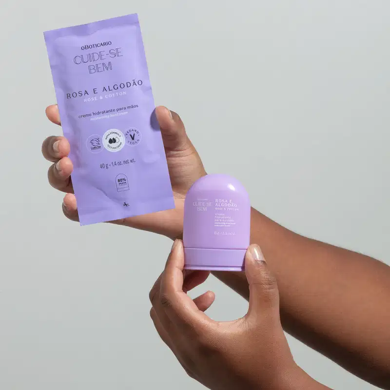 Uma mão segurando um refil de creme hidratante para mãos da marca Cuide-se Bem Rosa e Algodão em sachê de 40g na cor lilás com letras pretas e a outra segurando a embalagem original do produto na forma arredondada e na cor azul. O fundo da imagem é azul.