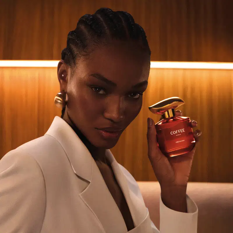 Imagem de uma mulher de pele negra segurando um frasco de desodorante colônia Coffee Woman Unique em um ambiente interno amadeirado com iluminação quente. Ela usa um casaco branco, brincos dourados e possui tranças no cabelo.
