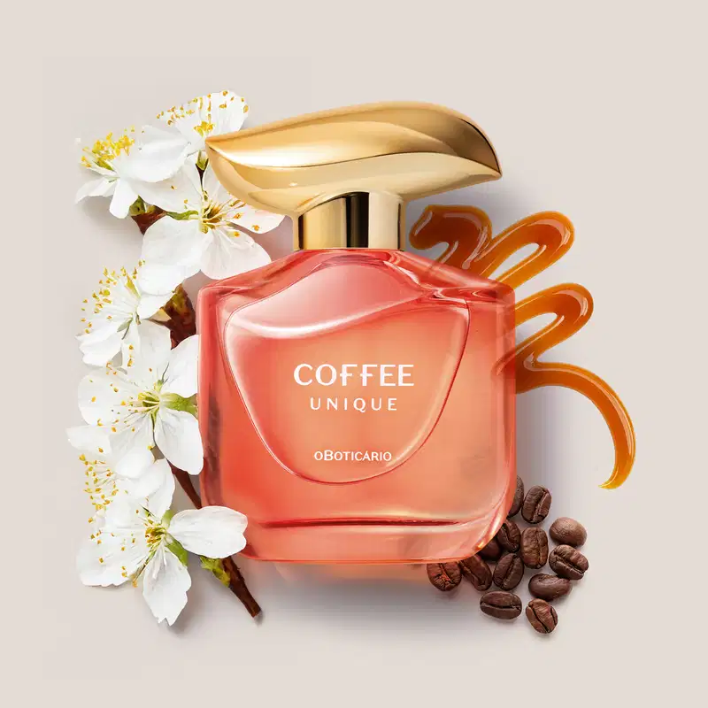 Imagem de um frasco rosa contendo 100ml de desodorante colônia da marca Coffee Woman Unique, com tampa dourada, em meio a flores brancas e grãos de café e um rastro de caramelo sobre superfície clara.