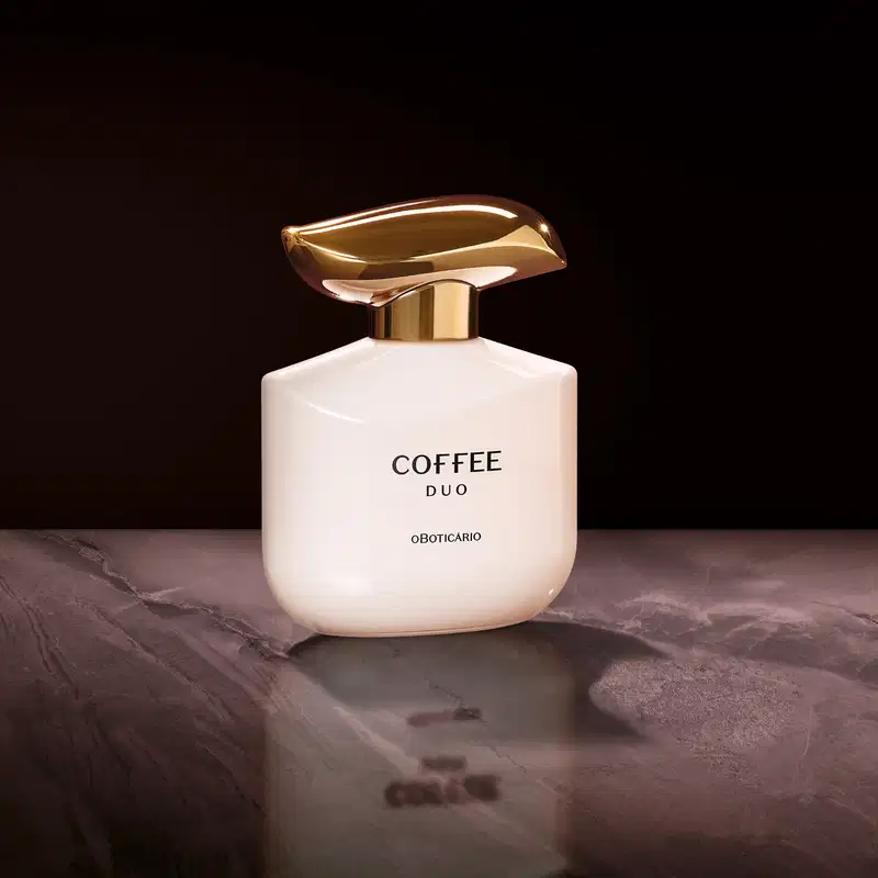 Imagem de um frasco do desodorante colônia Coffee Woman Duo em embalagem branca com tampa dourada em formato ondulado e rótulo em letras pretas. Ele está em um cenário com superfície iluminada e fundo escuro.