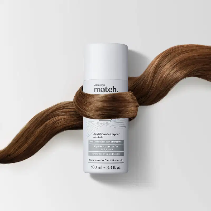 Um frasco branco com detalhes prateados e letras pretas e cinzas de acidificante capilar da marca Match Lab Tri Ação está em fundo branco e envolvido por uma mecha de cabelos sedosos e castanhos.