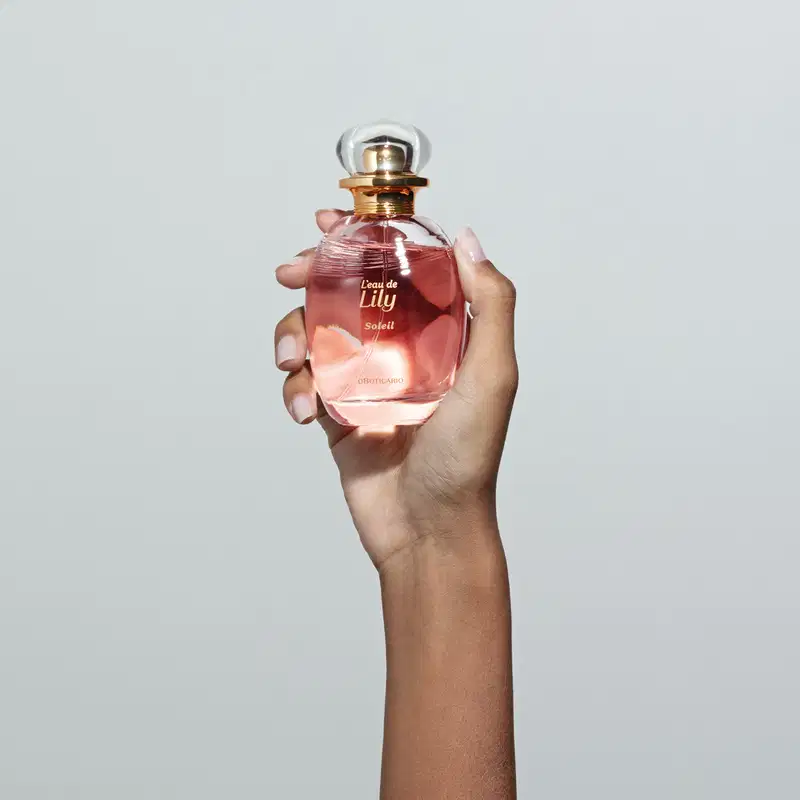 Mão feminina segurando um vidro transparente oval com líquido rosa, borrifador dourado e tampa transparente contendo desodorante colônia L'eau de Lily Soleil em fundo cinza.