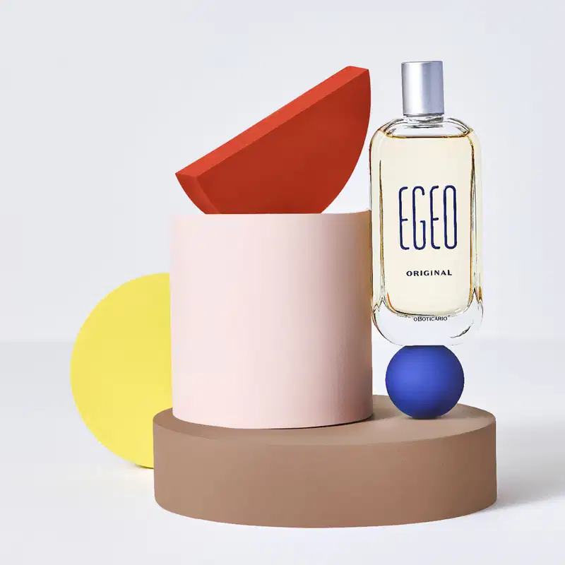 Vidro com líquido bege claro, tampa prateada e letras azul-marinho em relevo de desodorante colônia Egeo Original, posicionado em composição com cilindro rosa, formas geométricas em vermelho, amarelo e azul, sobre base circular bege em fundo cinza claro.