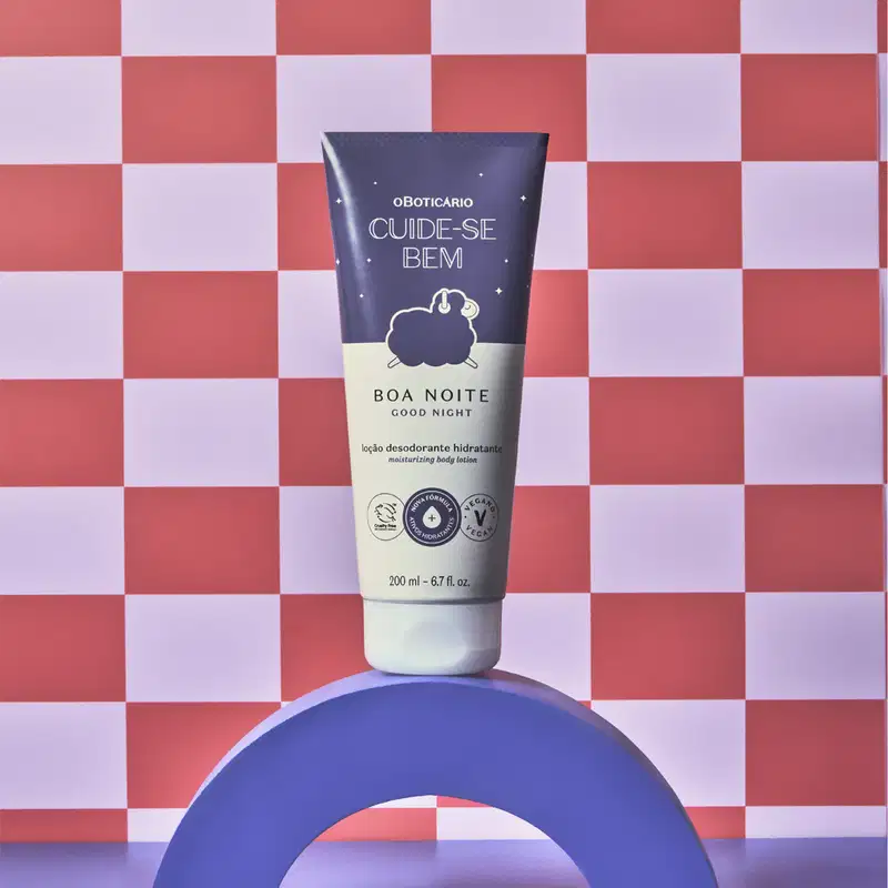 Bisnaga nas cores azul e lilás com tampa branca e ilustração de ovelhinha no rótulo contendo 200ml de loção hidratante corporal Cuide-se Bem Boa Noite em fundo xadrez vermelho e branco, apoiado sobre superfície azul.