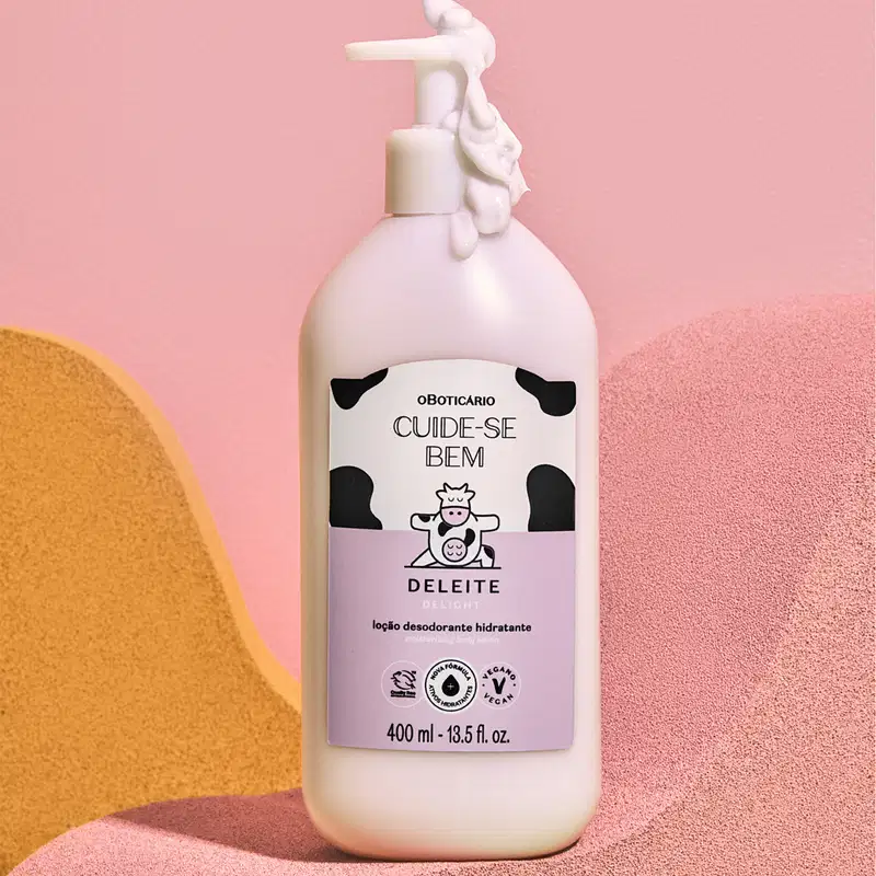 Frasco branco com bomba dosadora, rótulo branco e rosa com manchas pretas e com vaquinha ilustrada, de loção hidratante corporal Cuide-se Bem Deleite sobre superfície rosa e amarela com formas geométricas.