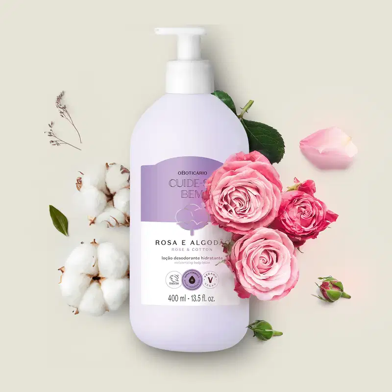 Frasco de 400ml na cor branca com rótulo lilás e branco com ilustração de uma flor de algodão contendo loção hidratante corporal Cuide-se Bem Rosa e Algodão posicionada entre flores de algodão e rosas cor de rosa em fundo bege claro.