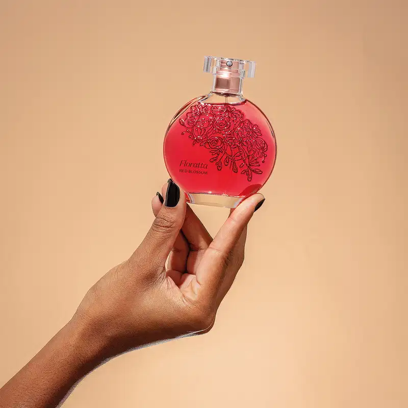 Mão feminina segurando um vidro transparente com líquido vermelho, florais em relevo, borrifador dourado e tampa cristal contendo desodorante colônia Floratta Red Blossom em fundo rosa.