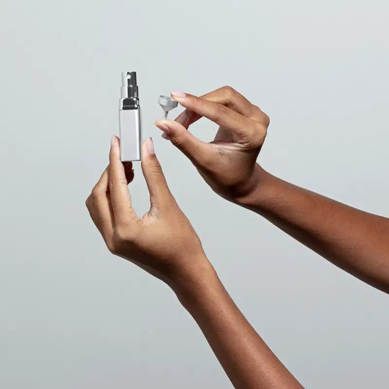 Uma mão feminina está segurando um porta-perfume com vaporizador em tubo prateado e a outra mao segura um mini funil em fundo azul claro.