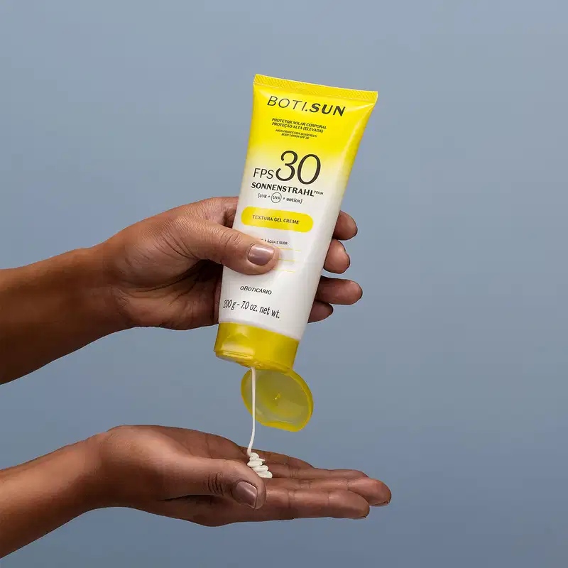 Mão segurando uma Bisnaga amarela e branca com tampa amarela e letras pretas contendo protetor solar corporal FPS 30 da marca Boti.Sun e jogando um pouco do creme branco na outra mão em fundo azul celeste.