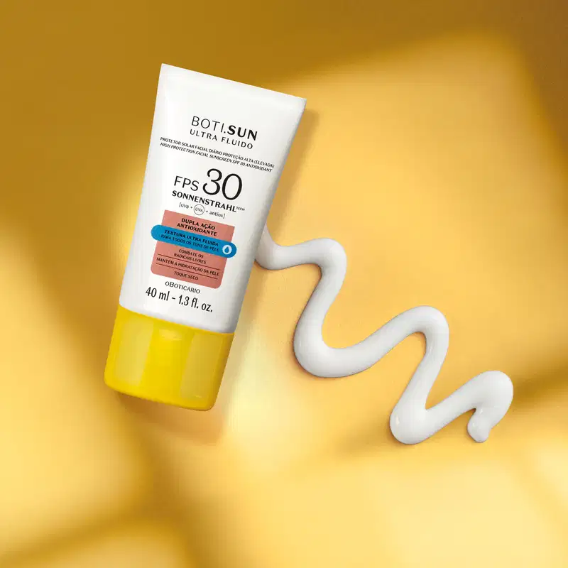 Frasco branco com tampa amarela, letras pretas e detalhes nas cores azul e laranja contendo protetor solar facial antioxidante FPS 30 Ultra Fluído da marca Boti.Sun em fundo laranja com sombra e uma pequena quantidade de creme branco ao lado.