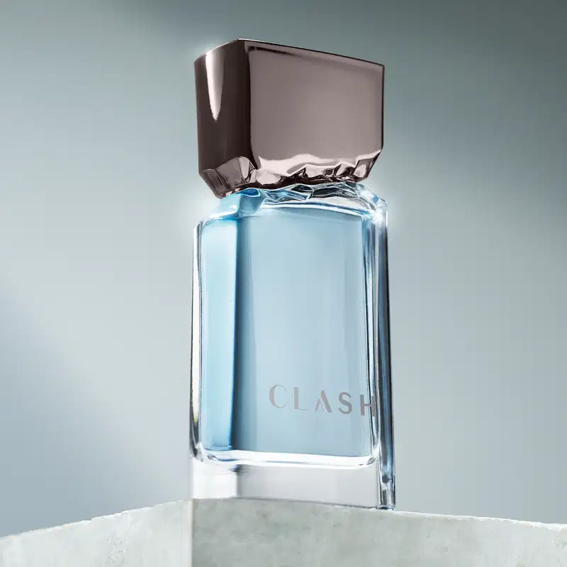 Frasco de item de perfumaria da marca Clash sobre superfície de mármore branco, com fundo desfocado em tons de azul e cinza.
