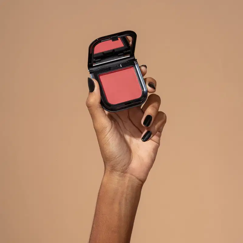 Uma mão feminina com unhas pintadas de preto está segurando um blush em pó mate Luminoso Berry Make B. na cor rosa intenso em estojo quadrado preto de cantos arredondados e espelho na tampa. O fundo da imagem é em tons de marrom.