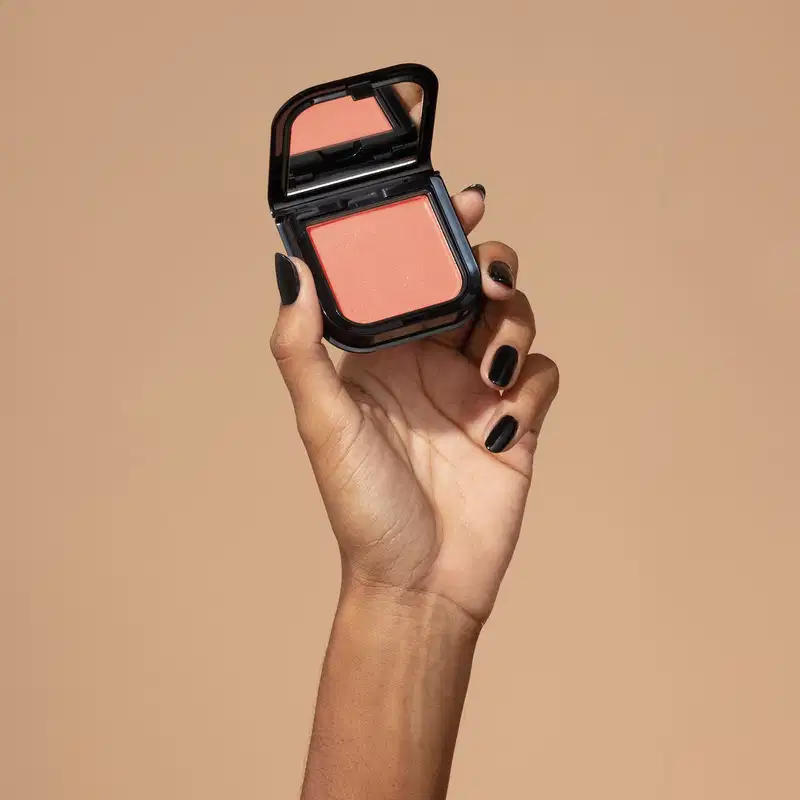 Uma mão feminina com unhas pintadas de preto está segurando um blush em pó mate Luminoso Coral Make B. na cor coral em estojo quadrado preto de cantos arredondados e espelho na tampa. O fundo da imagem é em tons de marrom.