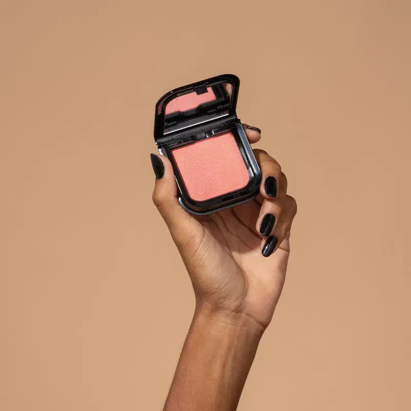 Uma mão feminina com unhas pintadas de preto está segurando um blush em pó mate Luminoso Rose Make B. na cor rosa em estojo quadrado preto de cantos arredondados e espelho na tampa. O fundo da imagem é em tons de marrom.