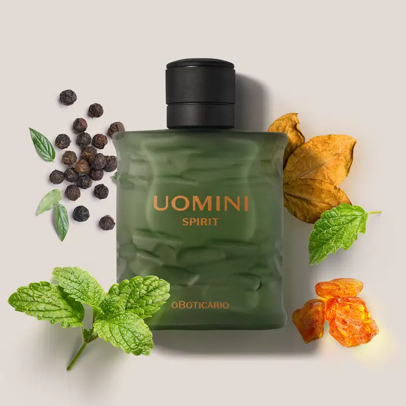 Frasco retangular em tom de verde oliva com tampa texturizada preta e letras laranjas de desodorante colônia Uomini Spirit., cercado por folhas verdes, pimenta-do-reino preta e laranja desidratada sobre superfície clara.