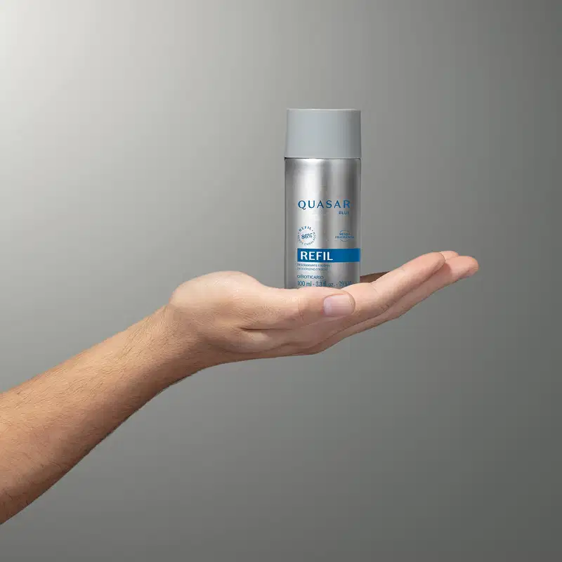 Mão segurando um refil de desodorante colônia Quasar Blue em frasco prateado com rótulo azul em fundo em tons de cinza.