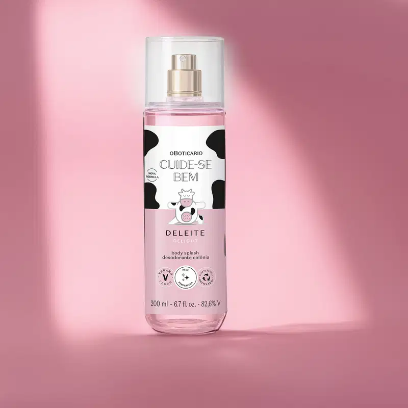 Frasco transparente com líquido rosa, pulverizador prateado, tampa transparente e rótulo branco e rosa com manchas pretas e ilustração de vaca contendo 200ml de body splash Cuide-se Bem Deleite em fundo rosa com sombras.
