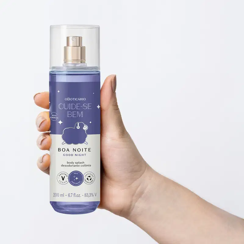 Mão segurando um Frasco com líquido azul claro, pulverizador prateado, tampa transparente e rótulo azul escuro e branco com ovelhinha ilustrada de body splash da marca Cuide-se Bem Boa Noite em fundo azul claro.