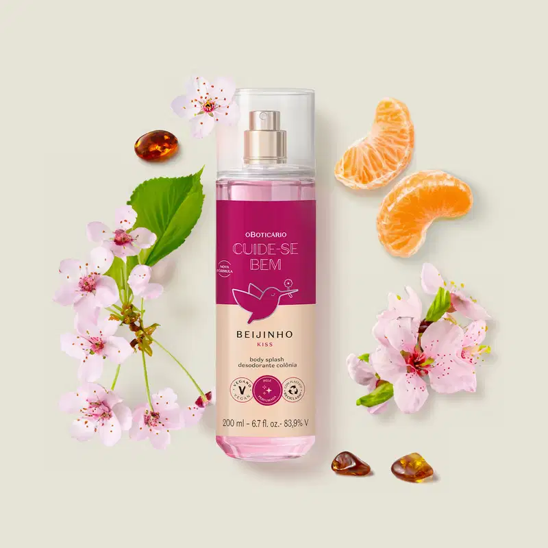 Frasco com líquido rosa, pulverizador prateado, tampa transparente e rótulo vinho e rosa com beija-flor ilustrado de body splash da marca Cuide-se Bem Beijinho. em ambiente com flores de cerejeira, fatias de laranja e pedras âmbar, sobre superfície clara.