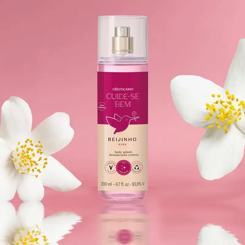 Frasco com líquido rosa, pulverizador prateado, tampa transparente e rótulo vinho e rosa com beija-flor ilustrado de body splash da marca Cuide-se Bem Beijinho em fundo rosa, com flores brancas ao redor, refletido em superfície brilhante.