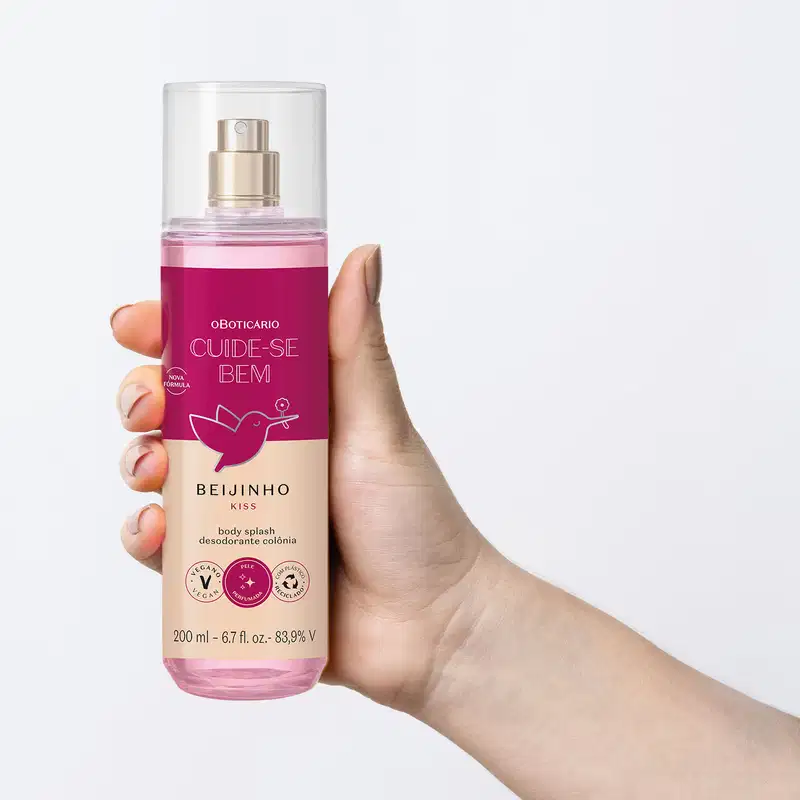 Mão segurando um frasco com líquido rosa, pulverizador prateado, tampa transparente e rótulo vinho e rosa com beija-flor ilustrado de body splash da marca Cuide-se Bem Beijinho com fundo azul claro.