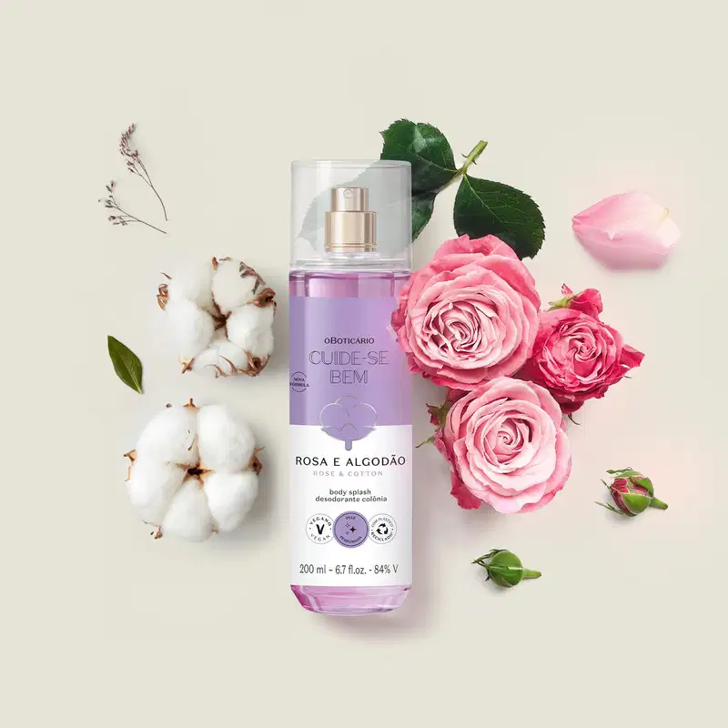 Frasco com líquido azul, pulverizador prateado, tampa transparente e rótulo azul e branco com flor de algodão ilustrada contendo body splash Cuide-se Bem Rosa e Algodão em meio a rosas cor de rosa, algodão e folhas verdes sobre superfície clara.