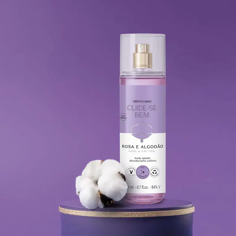 Frasco cilíndrico com líquido azul, pulverizador prateado, tampa transparente e rótulo azul e branco com flor de algodão ilustrada contendo body splash Cuide-se Bem Rosa e Algodão com flores de algodão ao redor em fundo azul escuro.