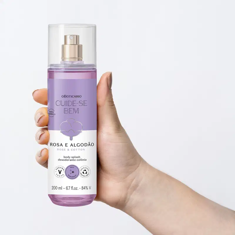Mão feminina segurando um frasco cilíndrico com líquido azul, pulverizador prateado, tampa transparente e rótulo azul e branco com flor de algodão ilustrada contendo body splash Cuide-se Bem Rosa e Algodão em fundo azul claro.