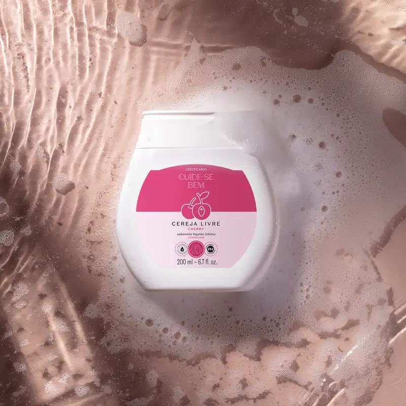 Recipiente branco com rótulo rosa da marca Cuide-se bem Cereja Livre, flutuando em espuma de banho com bolhas, em ambiente com iluminação suave e tons terrosos.
