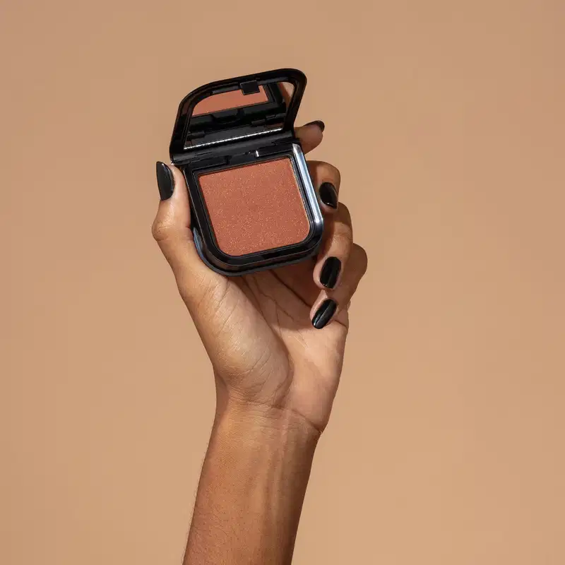 Uma mão feminina com unhas pintadas de preto está segurando um blush em pó mate Luminoso Brown Make B. na cor marrom em estojo quadrado preto de cantos arredondados e espelho na tampa. O fundo da imagem é em tons de marrom.