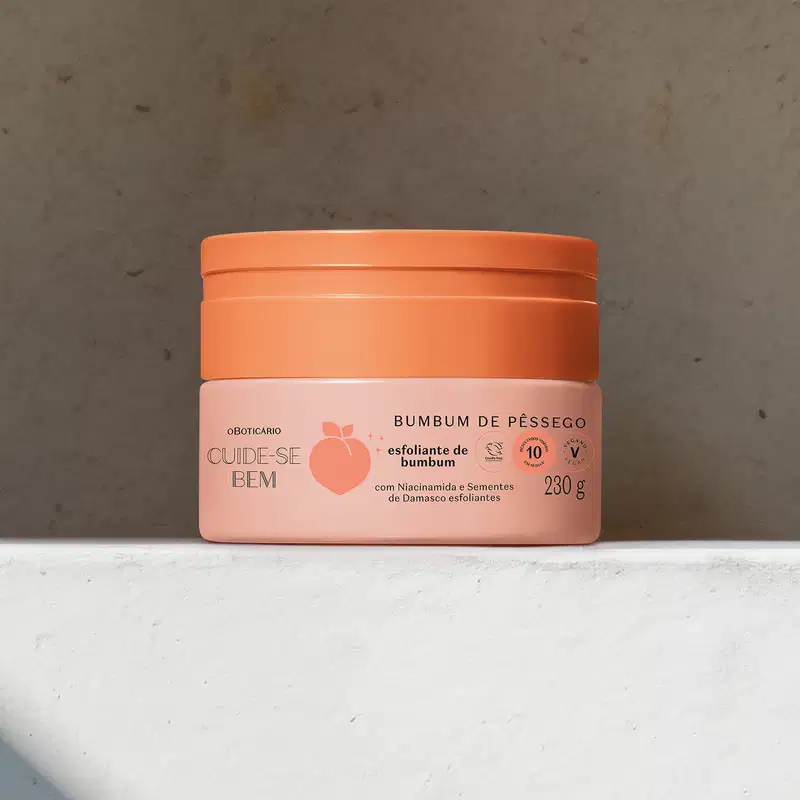 Pote em tons de rosa com letras pretas e pêssego ilustrado contendo creme esfoliante de bumbum Cuide-se Bem Bumbum de Pêssego em superfície clara e fundo marrom.