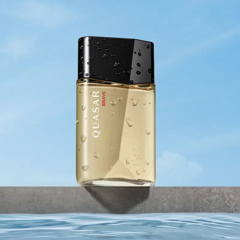 Frasco de desodorante colônia da marca Quasar Brave sobre superfície cinza, com gotas d'água, em cenário com céu azul e reflexo na água.