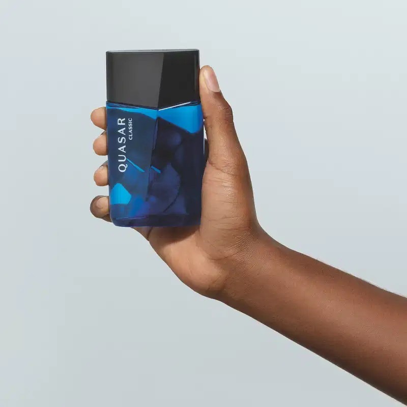 Mão masculina de pele negra segurando um vidro azul com tampa preta, ambos em formato geométrico e letras brancas no rótulo contendo 100ml de desodorante colônia Quasar Classic em fundo azul.