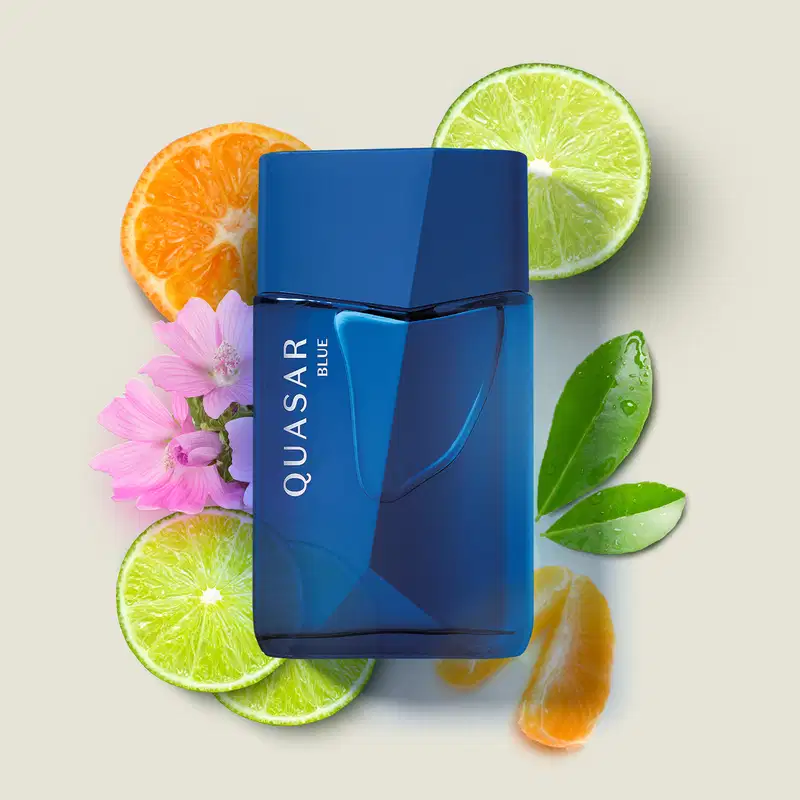 Frasco de vidro em formato geométrico com letras amarelas contendo desodorante colônia Quasar Blue sobre superfície clara, rodeado por fatias de frutas cítricas, folhas verdes e flores rosa.