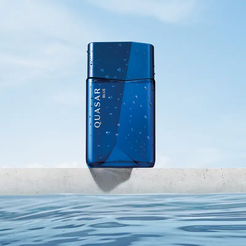 Frasco de vidro em formato geométrico com letras amarelas contendo desodorante colônia Quasar Blue com gotas d'água, flutuando sobre superfície de água em ambiente externo com céu claro.