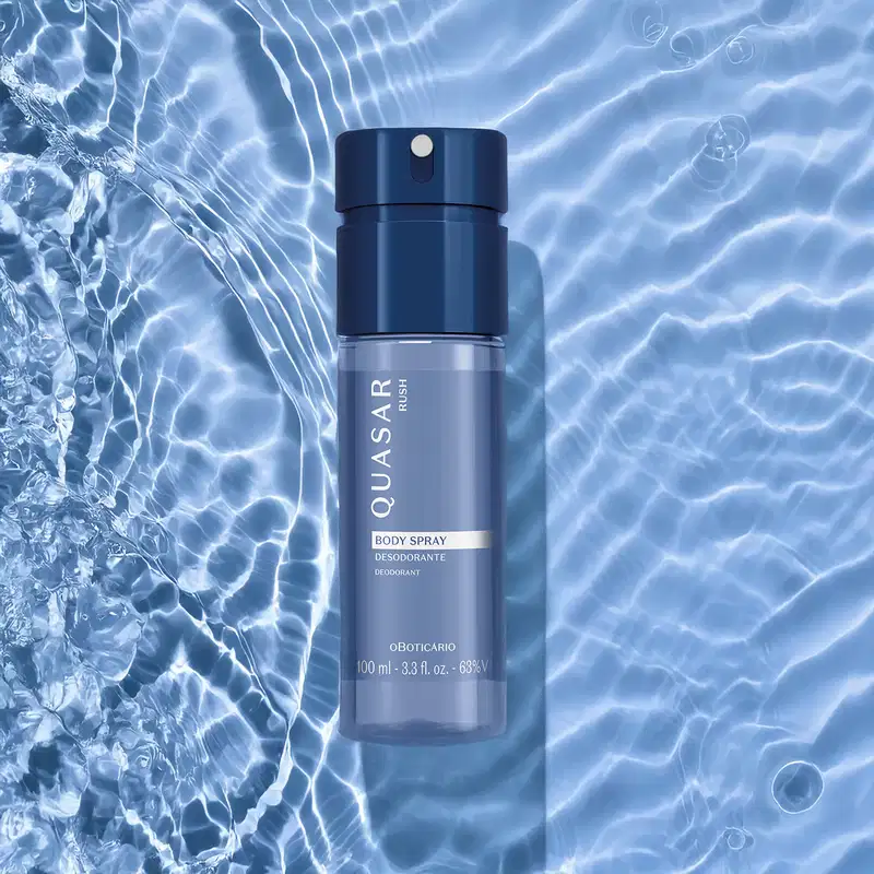 Frasco de body spray com líquido azul e tampa preta da marca Quasar Rush flutuando sobre superfície de água com ondulações e reflexos azulados.