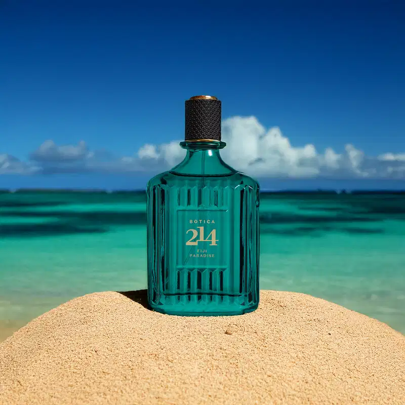 Vidro azul com textura ondulada, tampa preta e letras douradas de Eau de Parfum Fougére Especiado da marca Botica 214 Fiji Paradise. O produto está em um cenário de praia com areia, mar e céu azul.