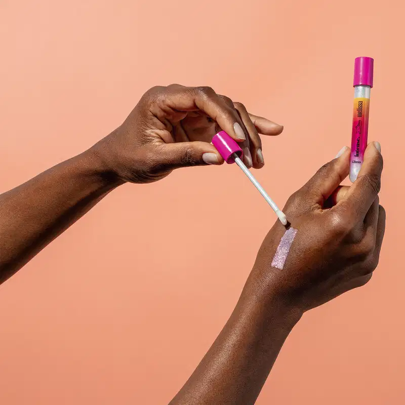 Uma mão feminina está segurando um tubo em tons de rosa, branco e laranja do gloss labial incolor Glossy Plastic Lips da marca Intense Melissa. A outra não está com a tampa e aplicador espalhando o produto na pele. O fundo da imagem é laranja.