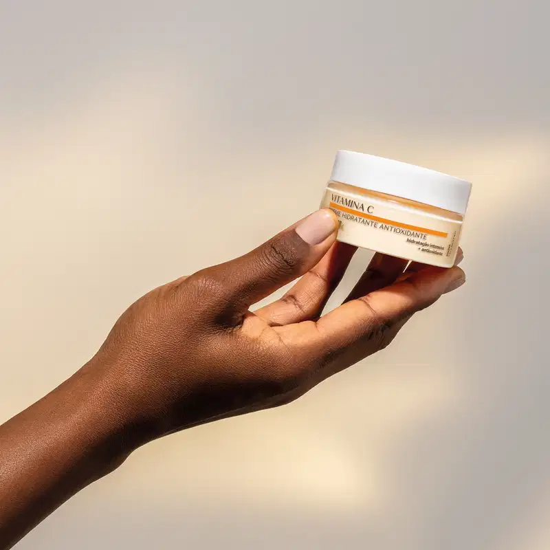 Mão feminina de pele negra segurando um pote de tampa branca, creme alaranjado e detalhe laranja de creme hidratante antioxidante Botik Vitamina C em fundo azul e branco.