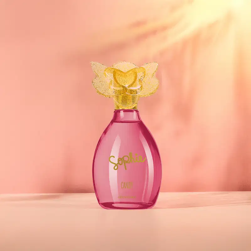 Frasco de colônia infantil Sophie Candy rosa com tampa dourada em formato de borboleta, sobre superfície clara em ambiente com iluminação suave e tons de laranja.s