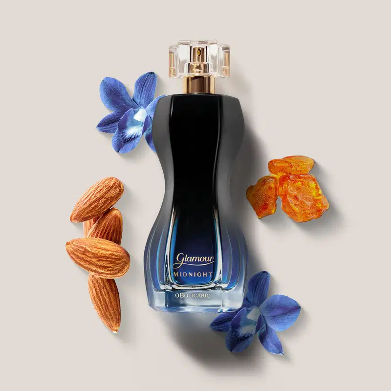 Vidro preto e azul de desodorante colônia Glamour Midnight rodeado por flores azuis, frutas secas alaranjadas e amêndoas sobre fundo bege. O cenário sugere um ambiente de luxo e sofisticação.
