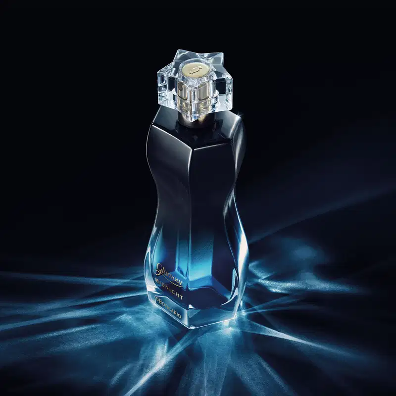 Vidro curvilíneo com degradê de preto e azul, borrifador dourado e tampa transparente de desodorante colônia Glamour Midnight em fundo escuro com reflexos e iluminação dramática, sugerindo um ambiente elegante e sofisticado.