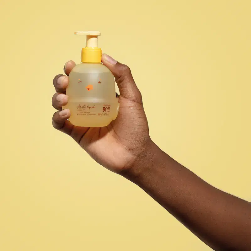 Mão segurando um frasco em forma de passarinho amarelo com detalhes laranjas contendo 200ml de sabonete líquido de glicerina da marca Boti Baby em um fundo alaranjado.