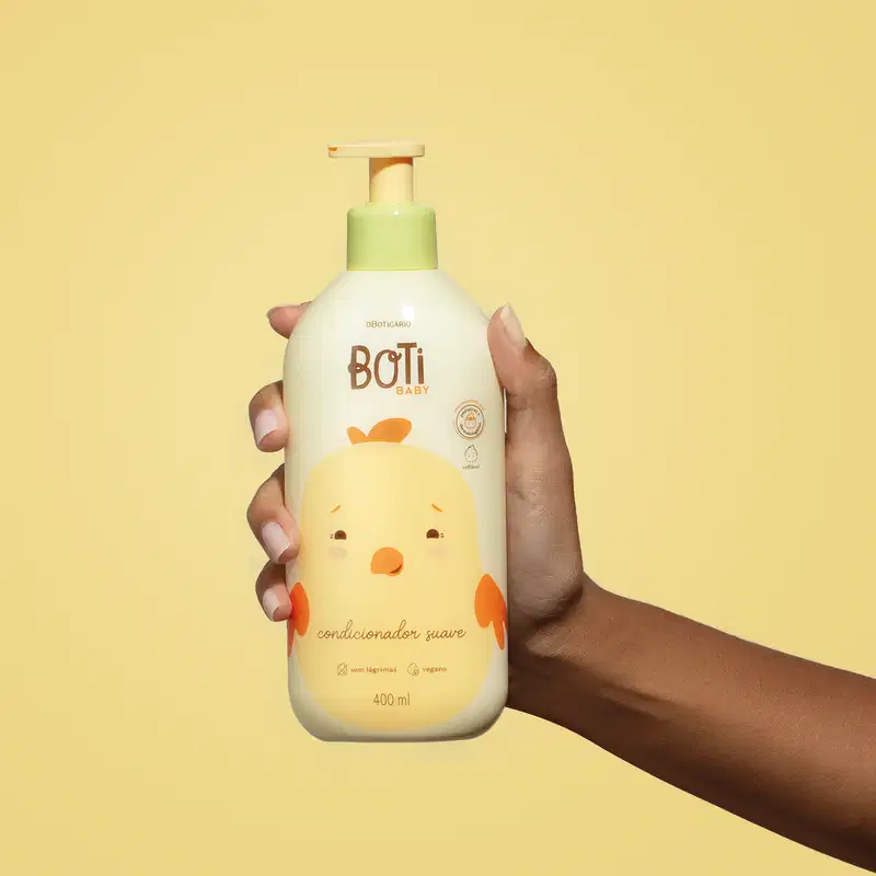 Mão segurando um frasco amarelo claro com ilustração de um passarinho amarelo com detalhes laranjas e bomba dosadora laranja de condicionador suave da marca Boti Baby em um fundo alaranjado.