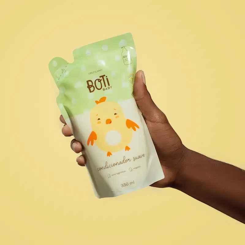 Mão segurando um refil de condicionador suave da marca Boti Baby em forma de sachê nas cores rosa e amarelo claro com ilustração de passarinho amarelo com detalhes laranjas em fundo alaranjado.