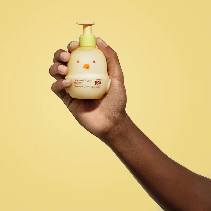 Mão segurando um frasco em forma de passarinho amarelo com detalhes laranjas contendo 200ml de condicionador suave da marca Boti Baby em um fundo alaranjado.