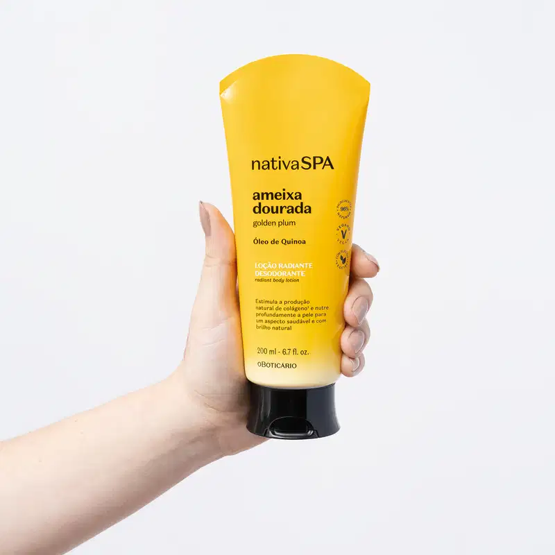 Mão segurando um frasco com degradê de amarelo e branco, tampa e letras pretas contendo loção radiante corporal Nativa SPA Ameixa Dourada em fundo azul claro.