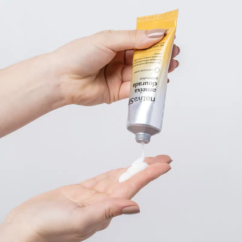 Uma mão está segurando uma bisnaga com degradê de prata e amarelo com letras pretas contendo gel creme para as mãos da marca Nativa SPA Ameixa Dourada e colocando um pouco do creme na outra mão. O fundo da imagem é azul claro.
