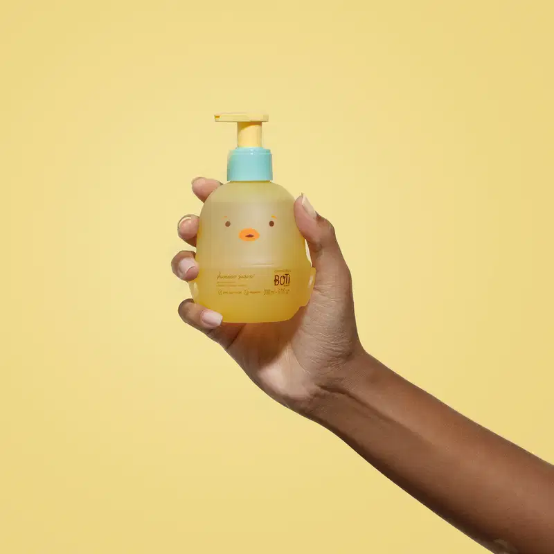 Mão segurando um frascoem forma de passarinho amarelo com detalhes laranjas, tampa azul e bomba dosadora amarela de shampoo suave da marca Boti Baby em um fundo alaranjado.