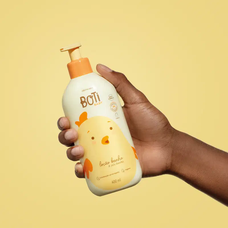 Mão segurando um frasco amarelo claro com ilustração de um passarinho amarelo com detalhes, tampa e bomba dosadora laranjas contendo 400ml de loção hidratante banho e pós banho da marca Boti Baby em um fundo alaranjado.